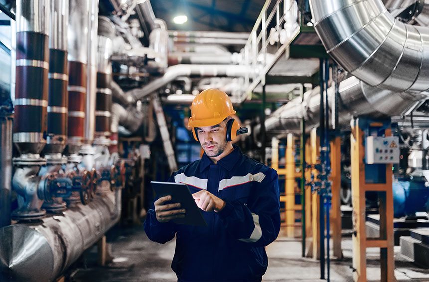 man wearing a hardhat working on a tablet in a room with industrial equipment