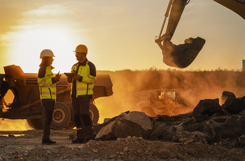 two workers in hardhats and high visibility vests talking with heavy machinery in the background